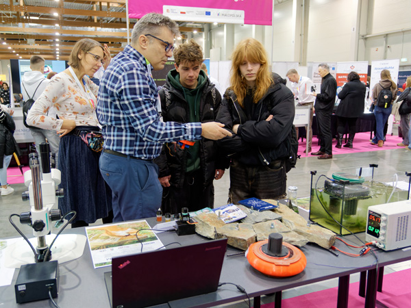 A man in a checked shirt points to a table with measuring equipment. He explains something to two young people standing next to him