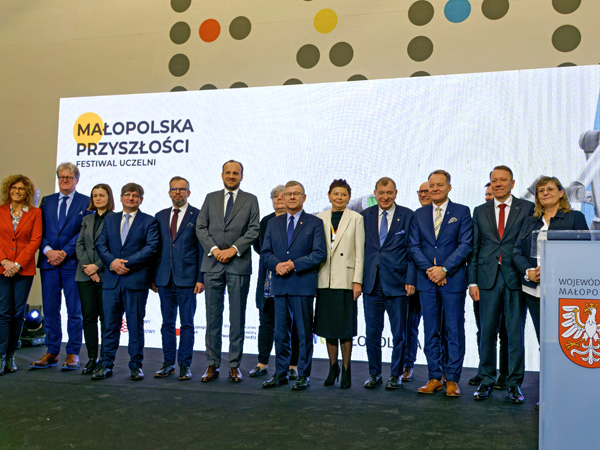 A group of elegantly dressed women and men stand in a row in front of a white screen displaying the festival logo