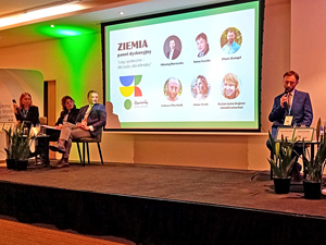 A screen showing a presentation. On either side are a man with a microphone and three people listening to him in chairs